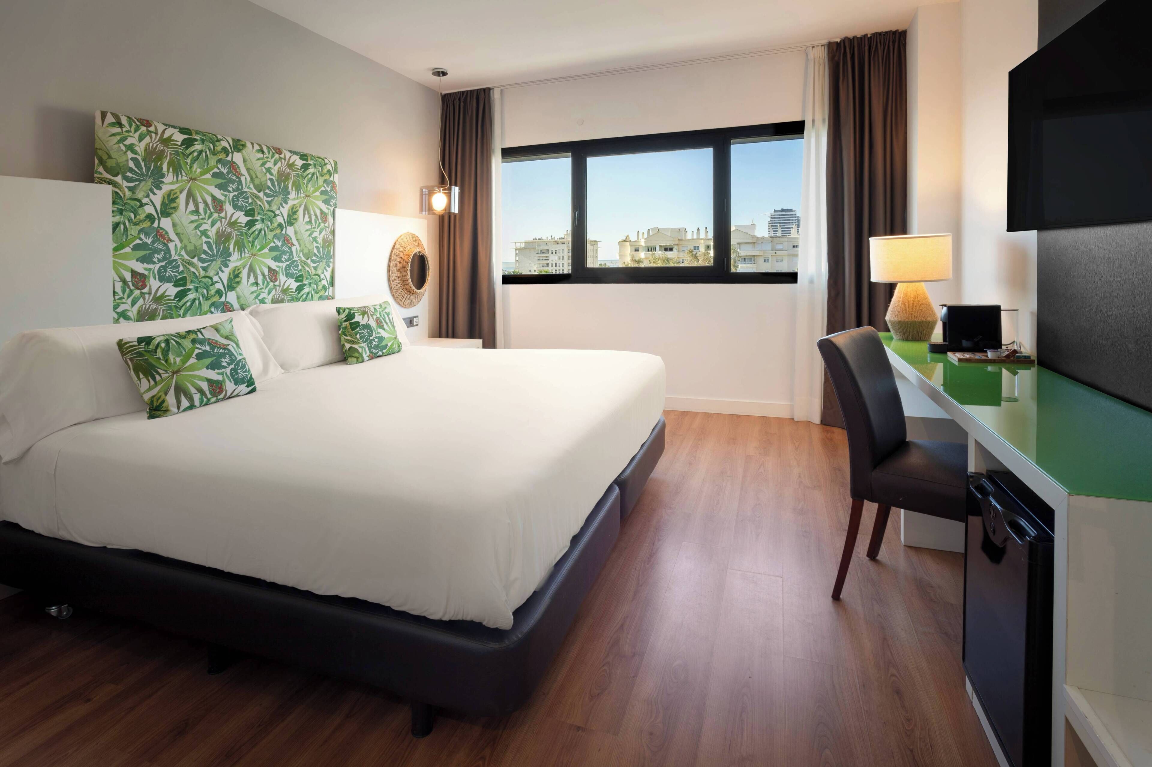Ein Hotelzimmer mit einem großen Bett, einem Schreibtisch mit Stuhl, einem Fernseher und einem Fenster mit Blick auf die Stadt.