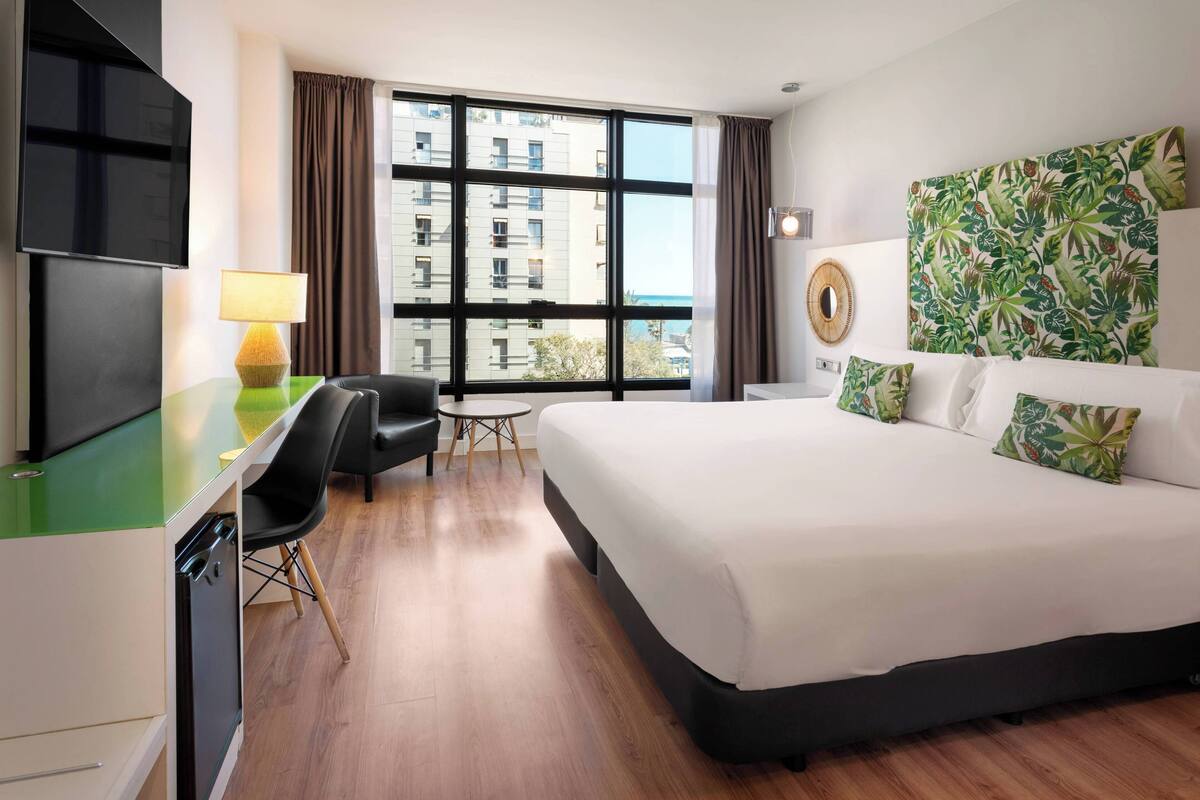 A hotel room with a large bed, a desk with a lamp, a chair, a TV, and a view of buildings through large windows.