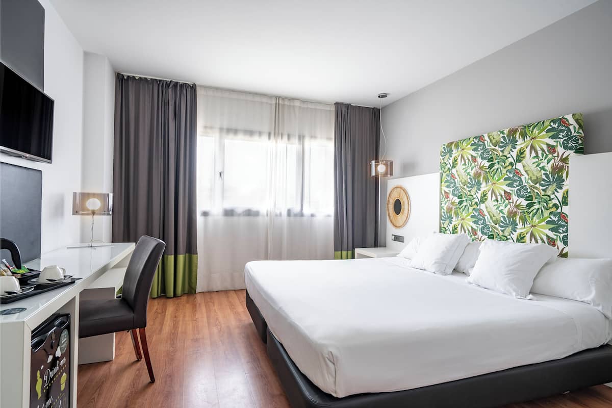 Une chambre d’hôtel comprenant un lit, un bureau avec une chaise, une télévision et un mur décoré d’un motif tropical.