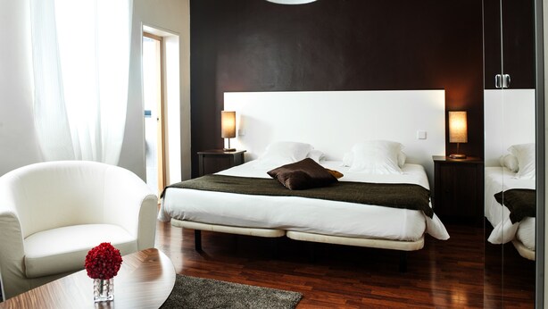 A modern hotel room with a large bed, two bedside tables with lamps, a white armchair, and a small table with a vase of flowers. - THC Gran Via Hostel (Madrid)
