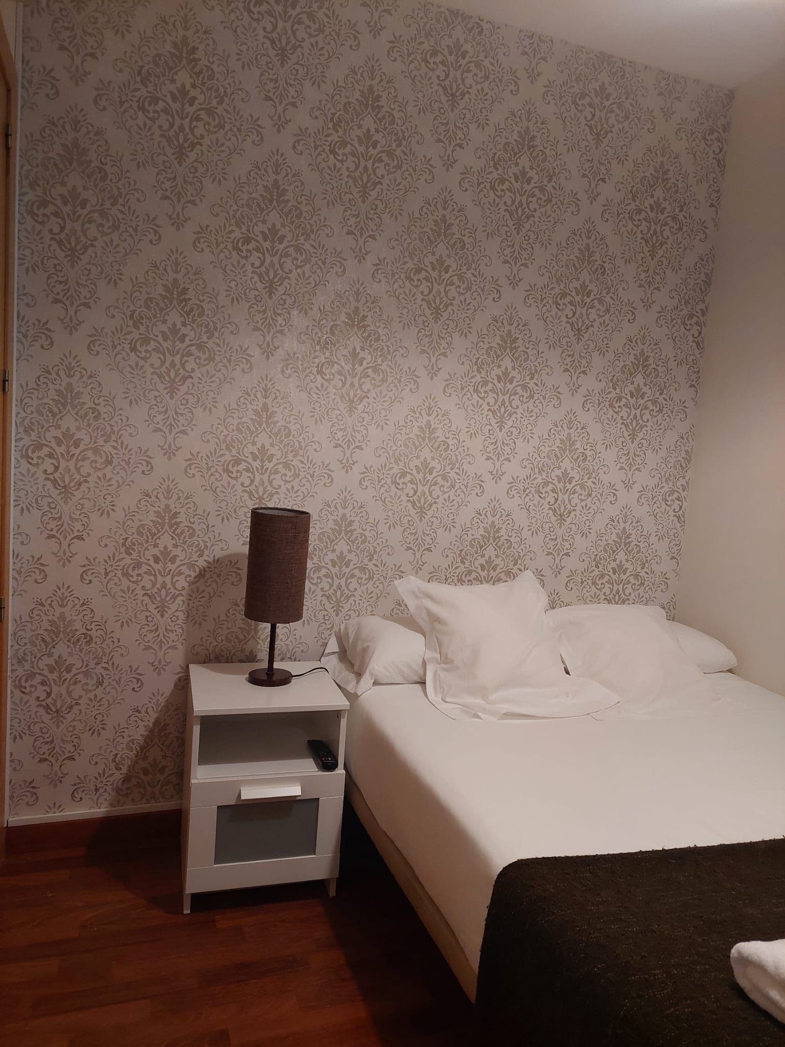 A bedroom with a bed, a nightstand, a lamp, and a patterned wallpaper.
