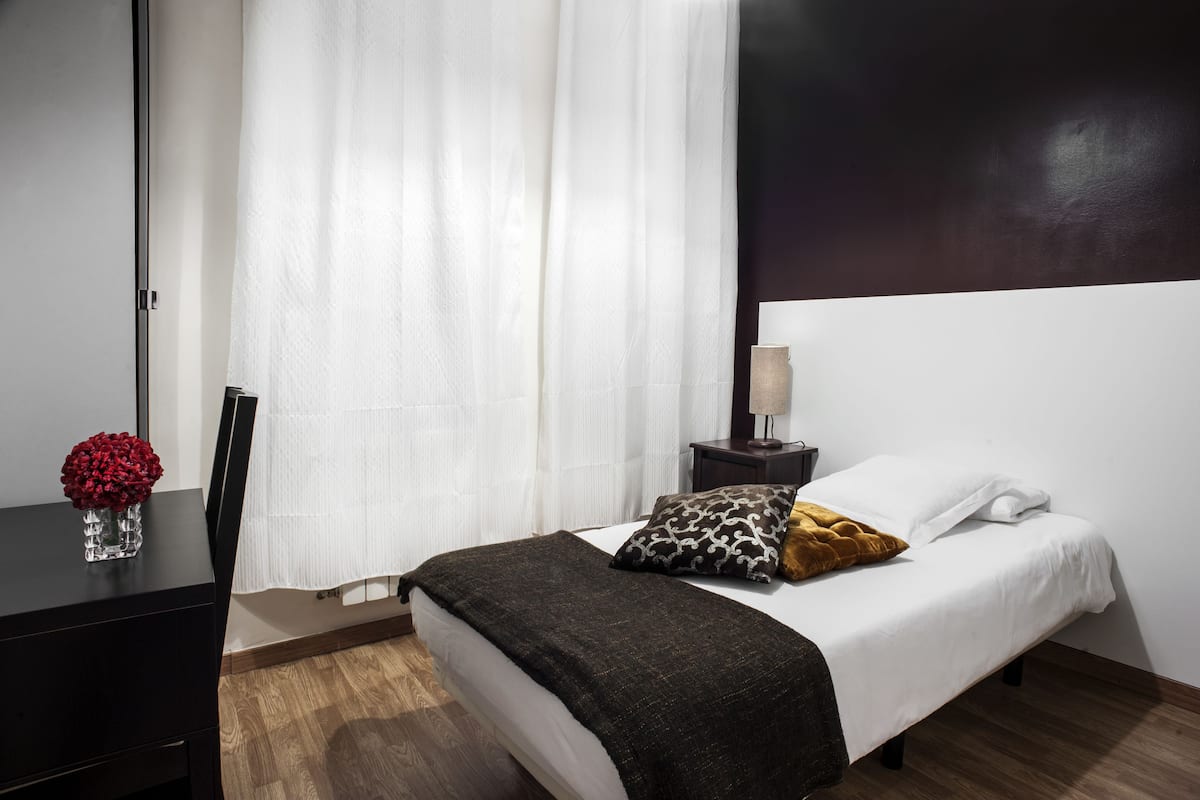 A hotel room with a bed, a nightstand, a lamp, a vase with flowers, and a window with curtains.