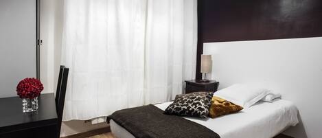 Habitación de hotel con cama, mesita de noche, lámpara, un florero con flores y una ventana con cortinas.