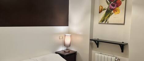 A hotel room with a bed, a nightstand with a lamp, a radiator, and a wall-mounted shelf with a painting.