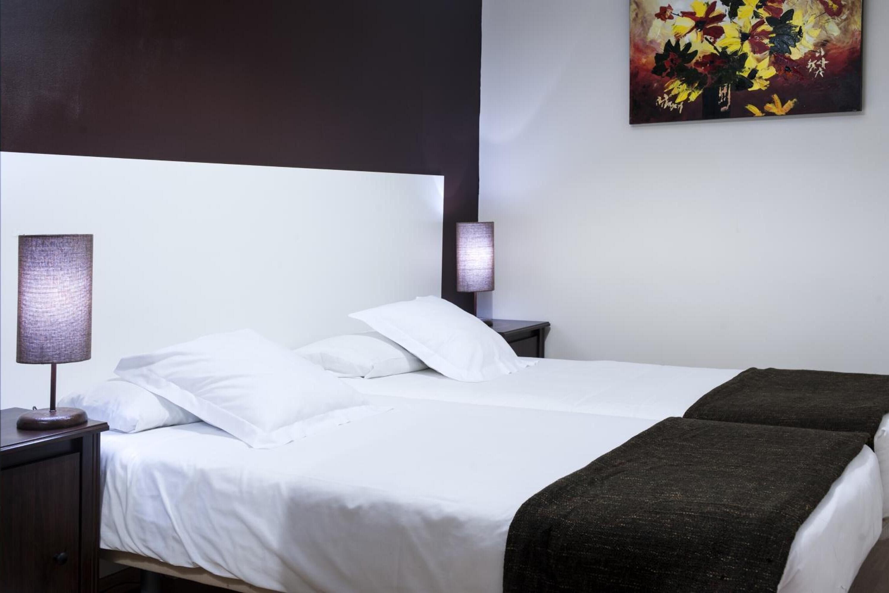 Ein ordentlich gemachtes Bett mit weißen Leinen und einer dunklen Tagesdecke, eine Nachttischlampe und eine Wandlampe.