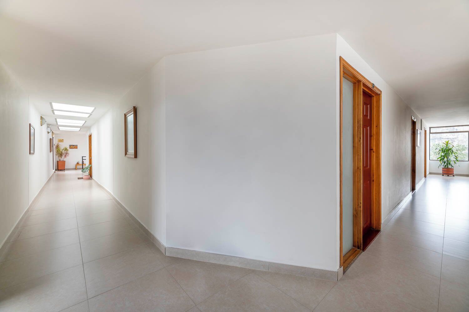 A long, narrow hallway with white walls and tiled flooring, leading to a wooden door and a window with a plant outside.