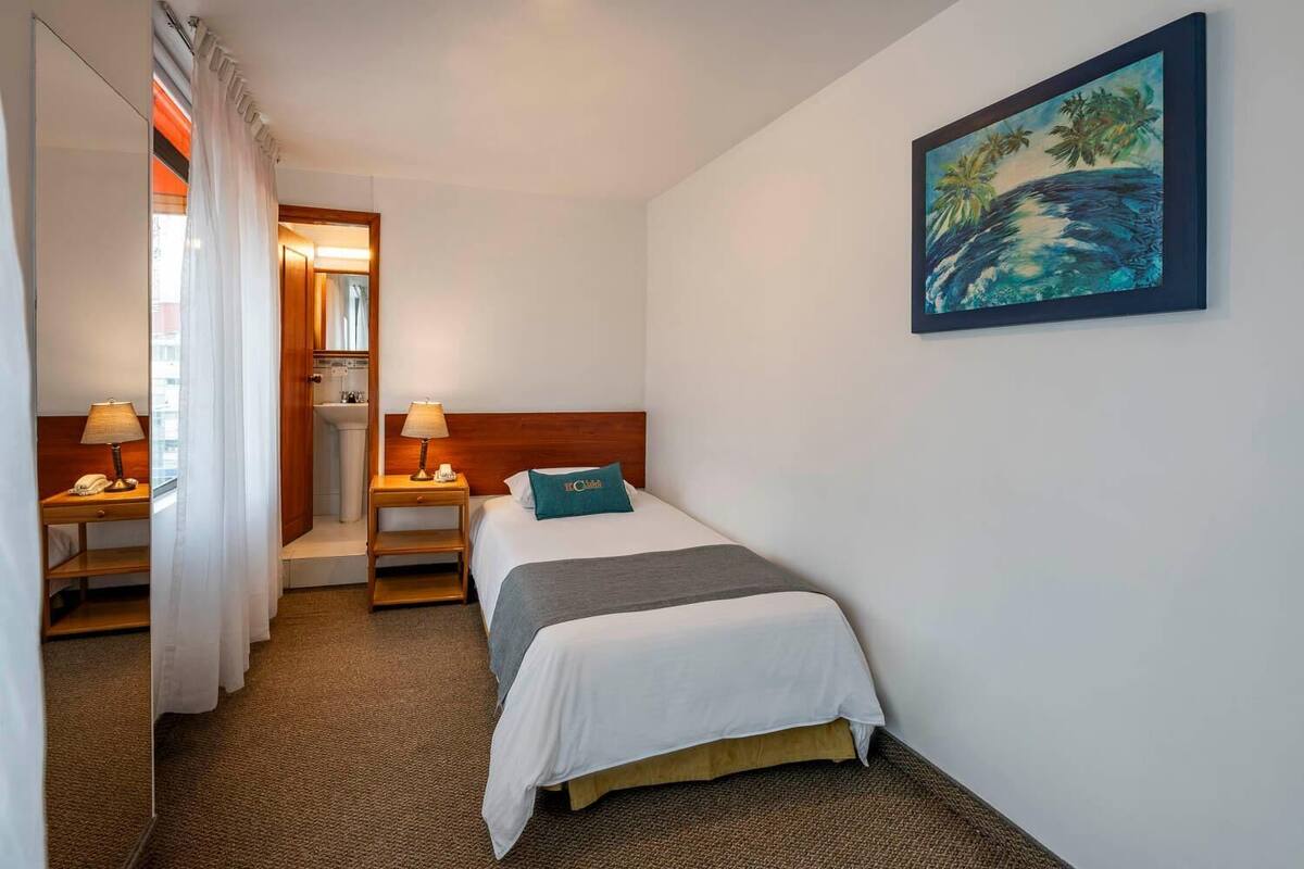A hotel room with a bed, a nightstand with a lamp, a bathroom with a sink, and a painting of a beach scene on the wall.