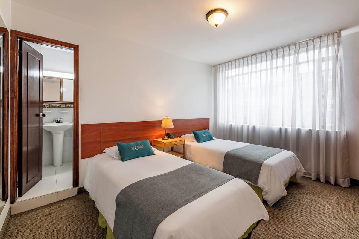 A hotel room with two beds, a nightstand with a lamp, a bathroom visible through an open door, and sheer curtains on the window.