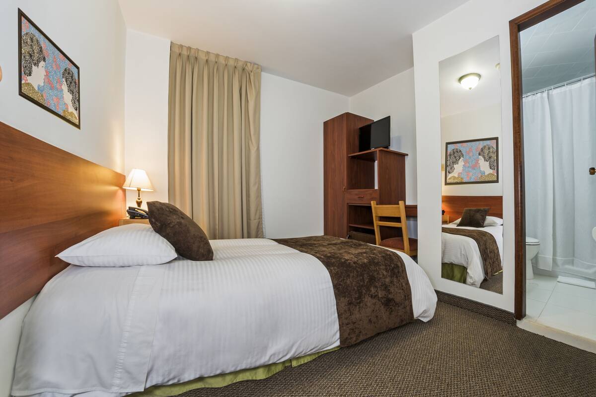 A hotel room with a large bed, a wooden headboard, a desk with a chair, a television, and a mirror.