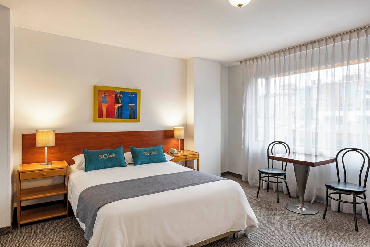 A hotel room with a bed, two bedside tables, a nightstand, a desk with two chairs, and a window with sheer curtains.
