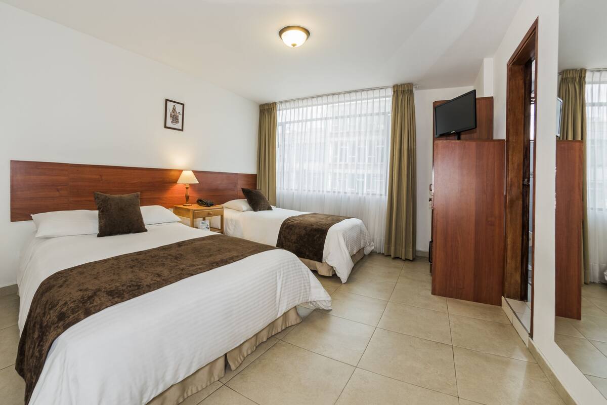 A hotel room with two beds, a wooden headboard, a bedside table with a lamp, a TV, and a window with curtains.