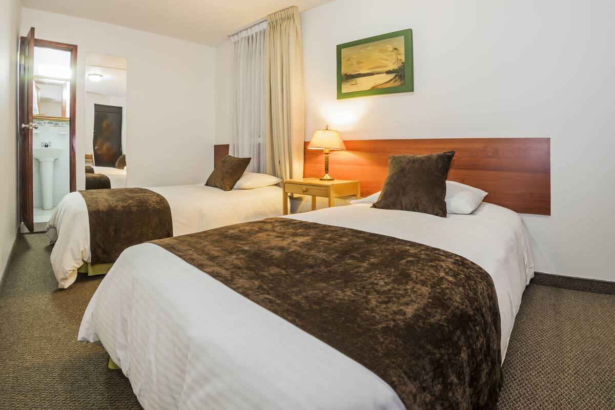 A hotel room with two beds, a bedside table with a lamp, a painting on the wall, and a bathroom visible through an open door.