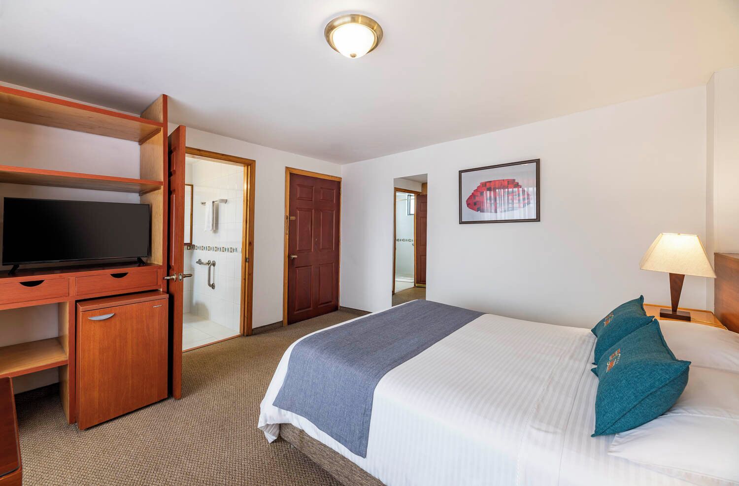 A hotel room with a bed, a desk with a TV, a door, and a framed picture on the wall.