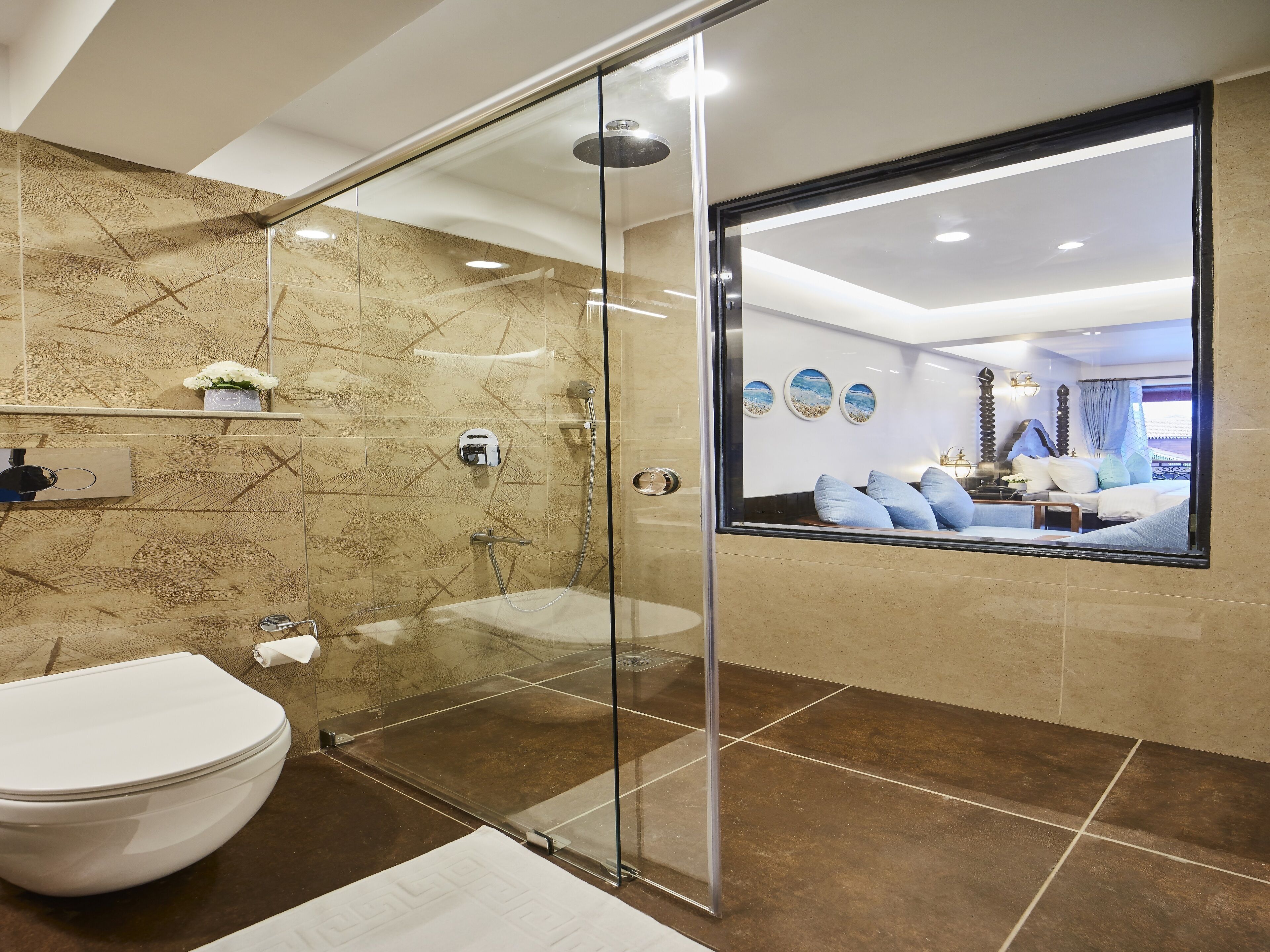 A modern bathroom with a glass shower enclosure, a toilet, and a large mirror reflecting a bedroom with a bed and decorative wall art.