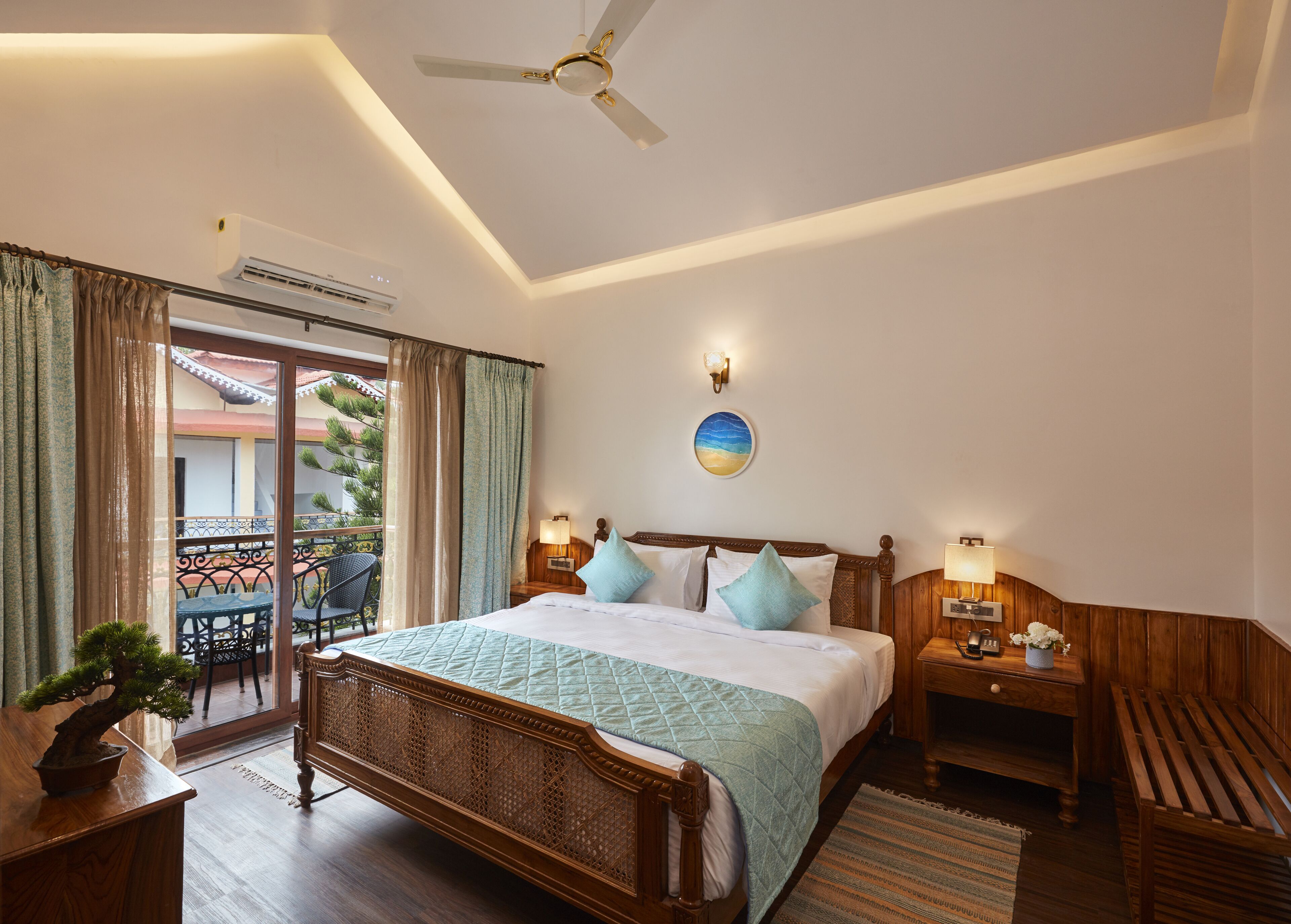 A hotel room with a bed, a ceiling fan, a nightstand, a wooden bench, a potted plant, and a balcony with outdoor furniture.