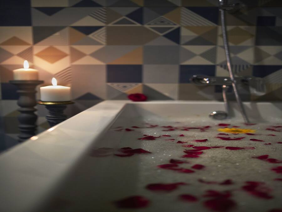 Un baño con una bañera llena de pétalos de rosa, dos velas encendidas y un revestimiento de azulejos geométricos.