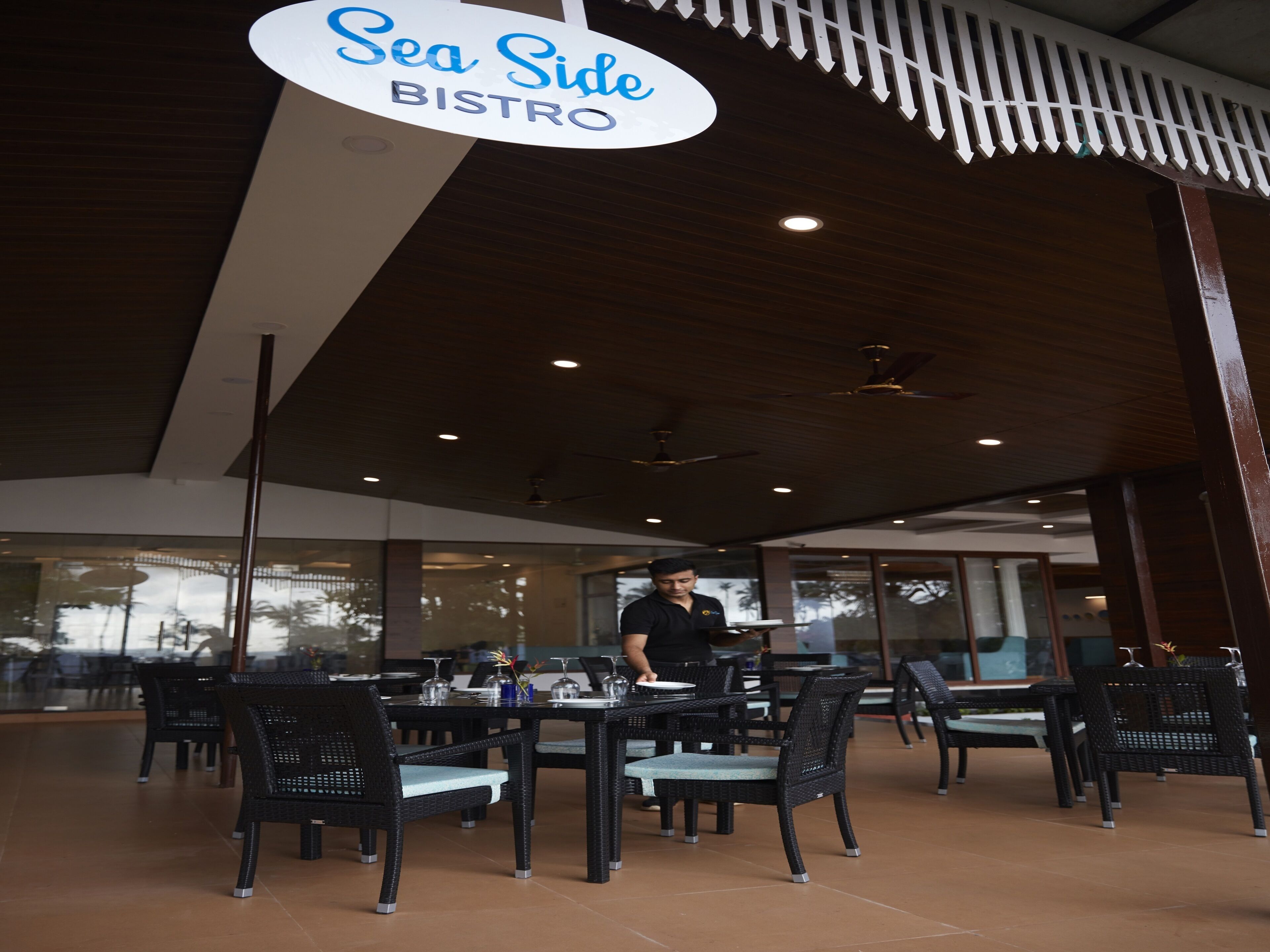 A restaurant interior with a sign reading "Sea Side BISTRO," featuring black wicker chairs, glass tables, and a modern ceiling design.