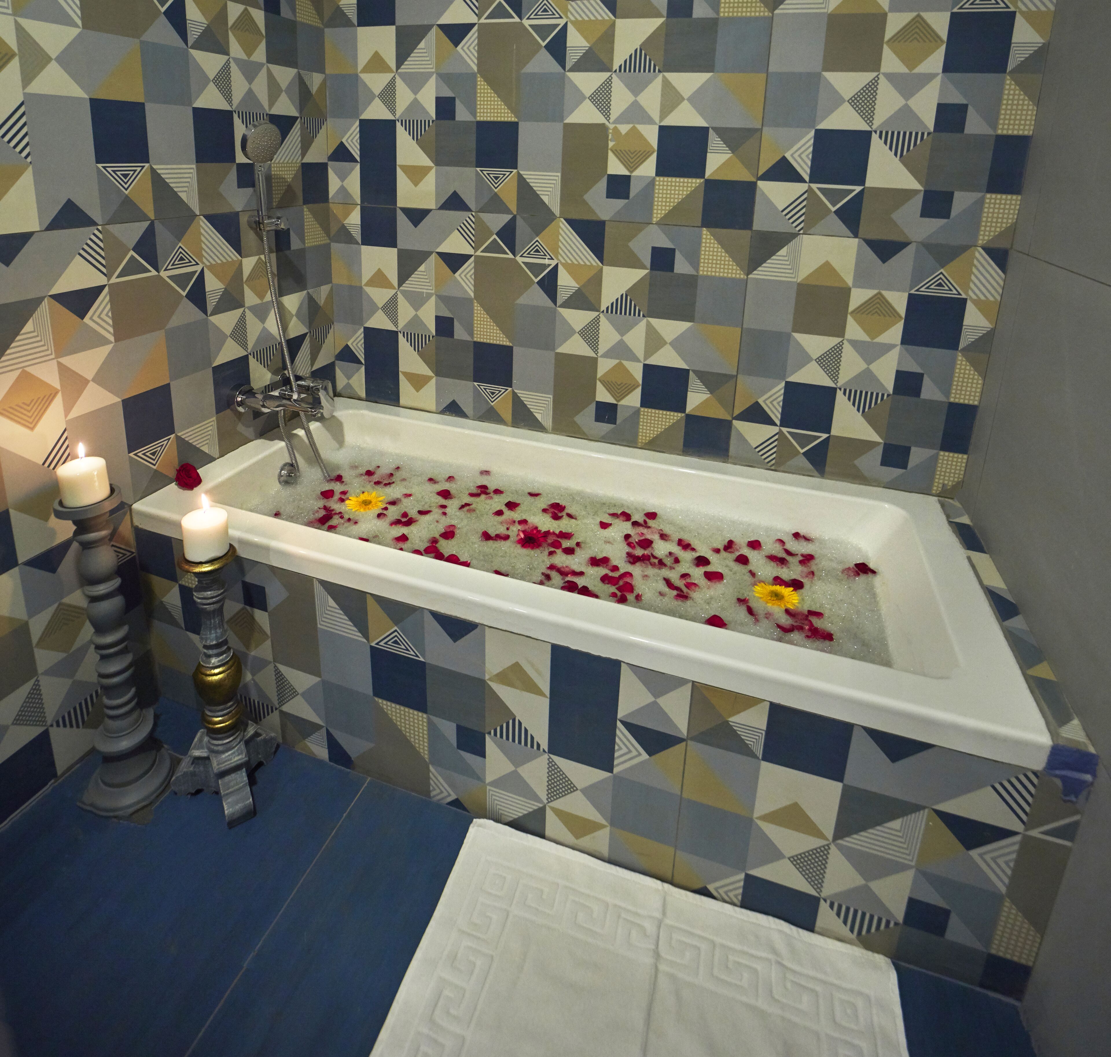 Een badkamer met een bad, geometrische tegels, twee kaarsen en rozenblaadjes in het water.