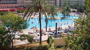 A large outdoor swimming pool surrounded by palm trees and lounge chairs, with a multi-story building in the background. - Club Cala Romani (Manacor)