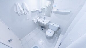 Un baño pequeño y blanco con inodoro, lavamanos y espejo. Hay toallas colgadas en la pared y un cubo de basura en el suelo.