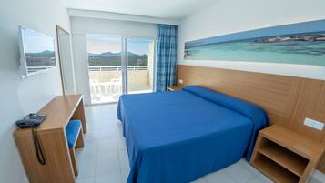 A hotel room with a bed, a desk, a chair, a small table, a painting of a beach, and a balcony with a view of the sea and mountains.