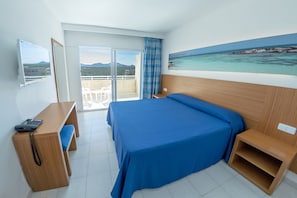 A hotel room with a bed, a desk, a chair, a small table, a painting of a beach, and a balcony with a view of the sea and mountains. - Club Cala Romani (Manacor)