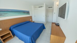 A hotel room with a bed, a desk, a chair, a TV, and a painting of a coastal scene. - Club Cala Romani (Manacor)