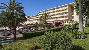 A hotel with multiple floors, a pool area with sun loungers, and palm trees. - Club Cala Romani (Manacor)