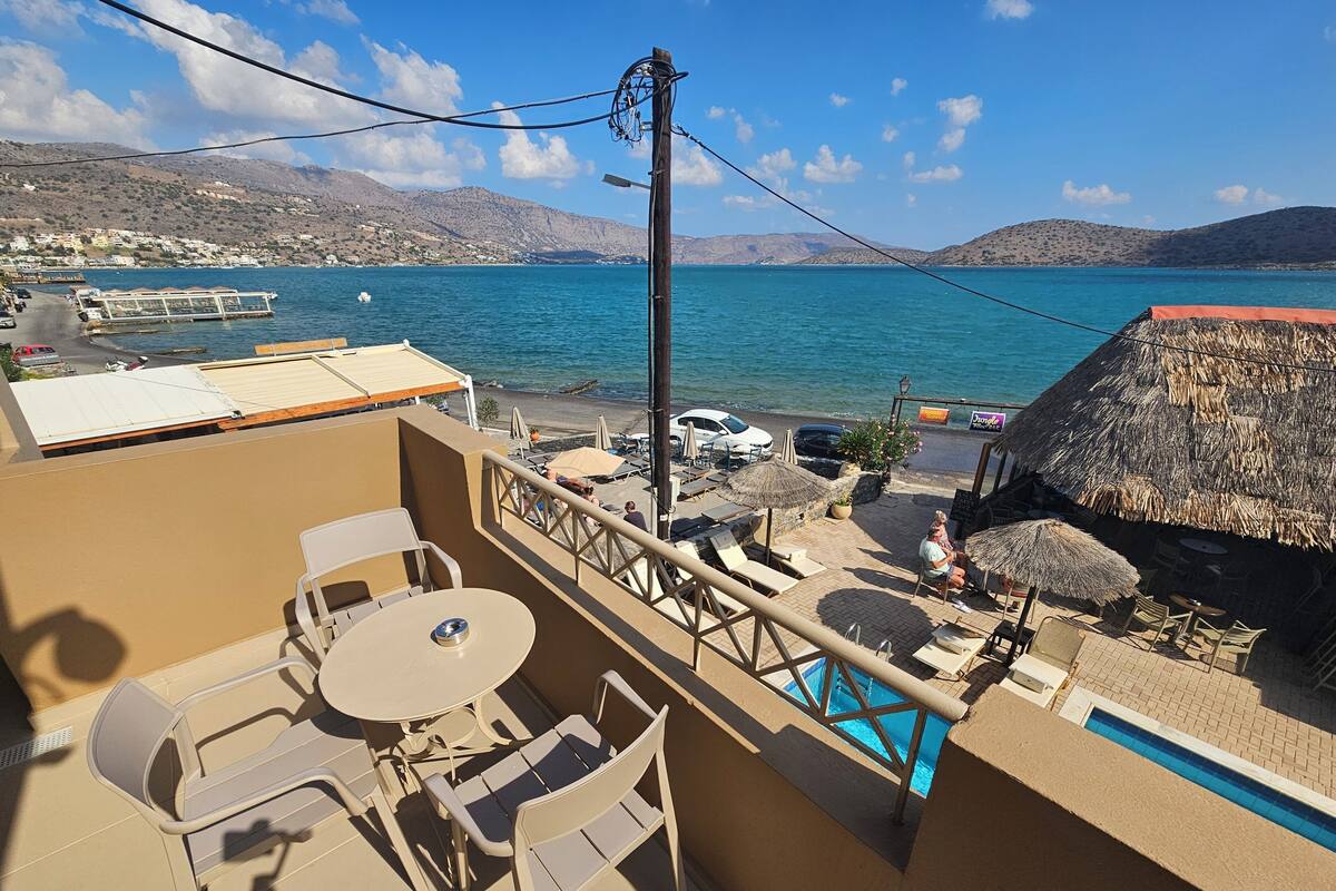 A balcony with a table and chairs, overlooking a beach with umbrellas and a pool.