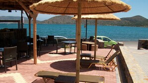 An outdoor seating area with tables and chairs, a thatched umbrella, and a view of the sea. - Elounda Sunrise Apartments (Agios Nikolaos)