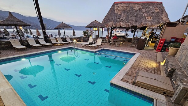 A swimming pool with lounge chairs and umbrellas, a bar area, and a view of the sea and mountains. - Elounda Sunrise Apartments (Agios Nikolaos)