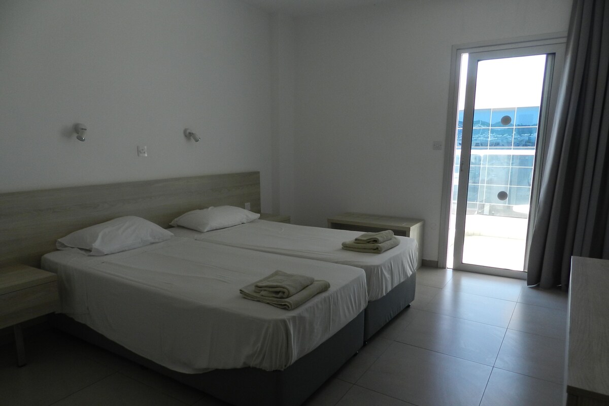 A hotel room with two beds, a small table, and a view of the city through a large window.