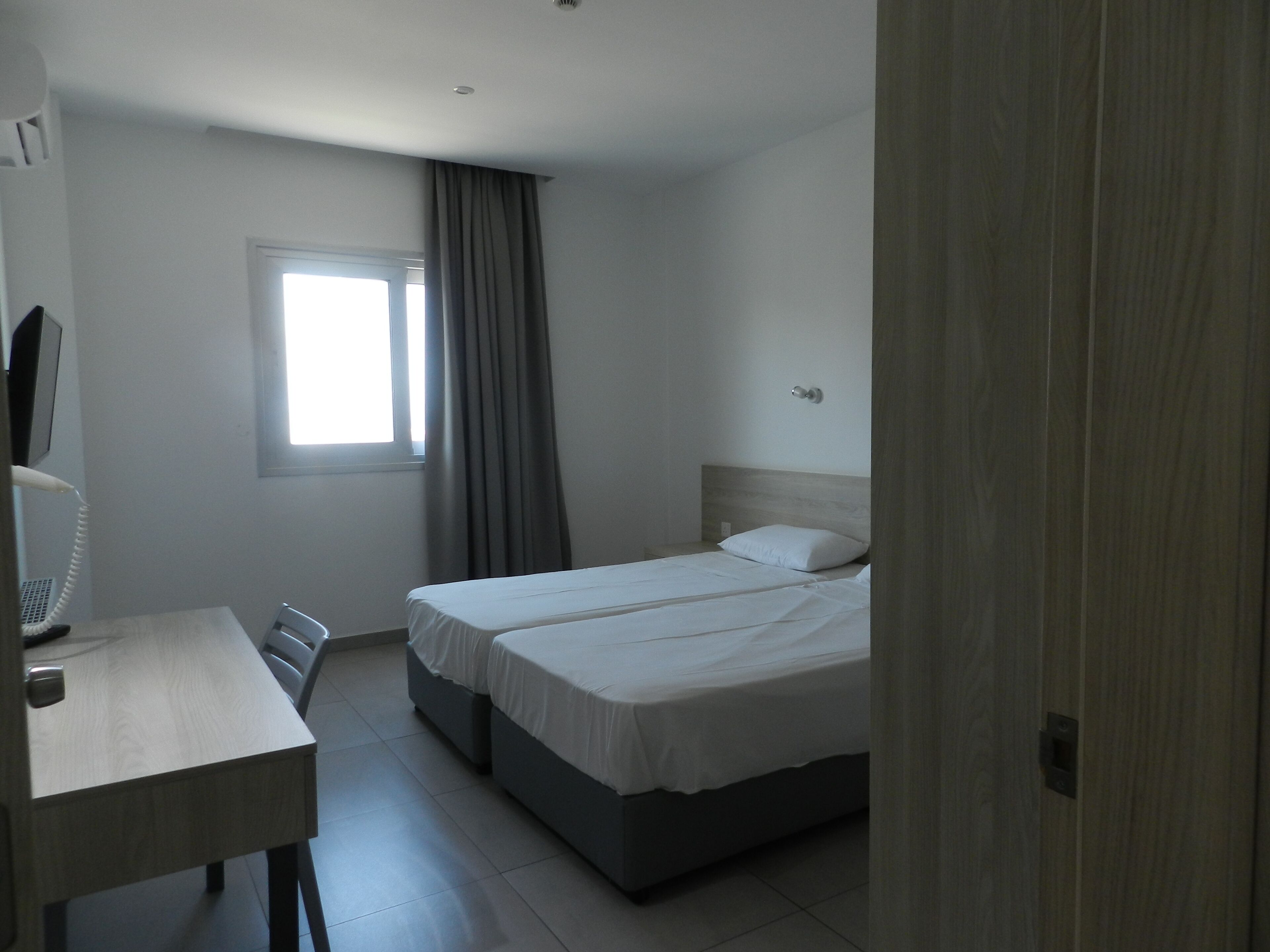 A hotel room with a bed, a desk, a chair, a window with curtains, and a wall-mounted phone.