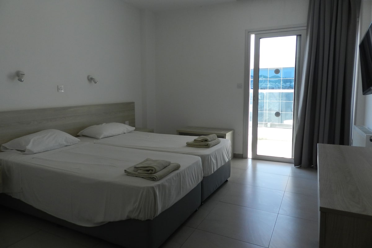 A hotel room with two beds, a small table, a window with curtains, and a view of the outside.