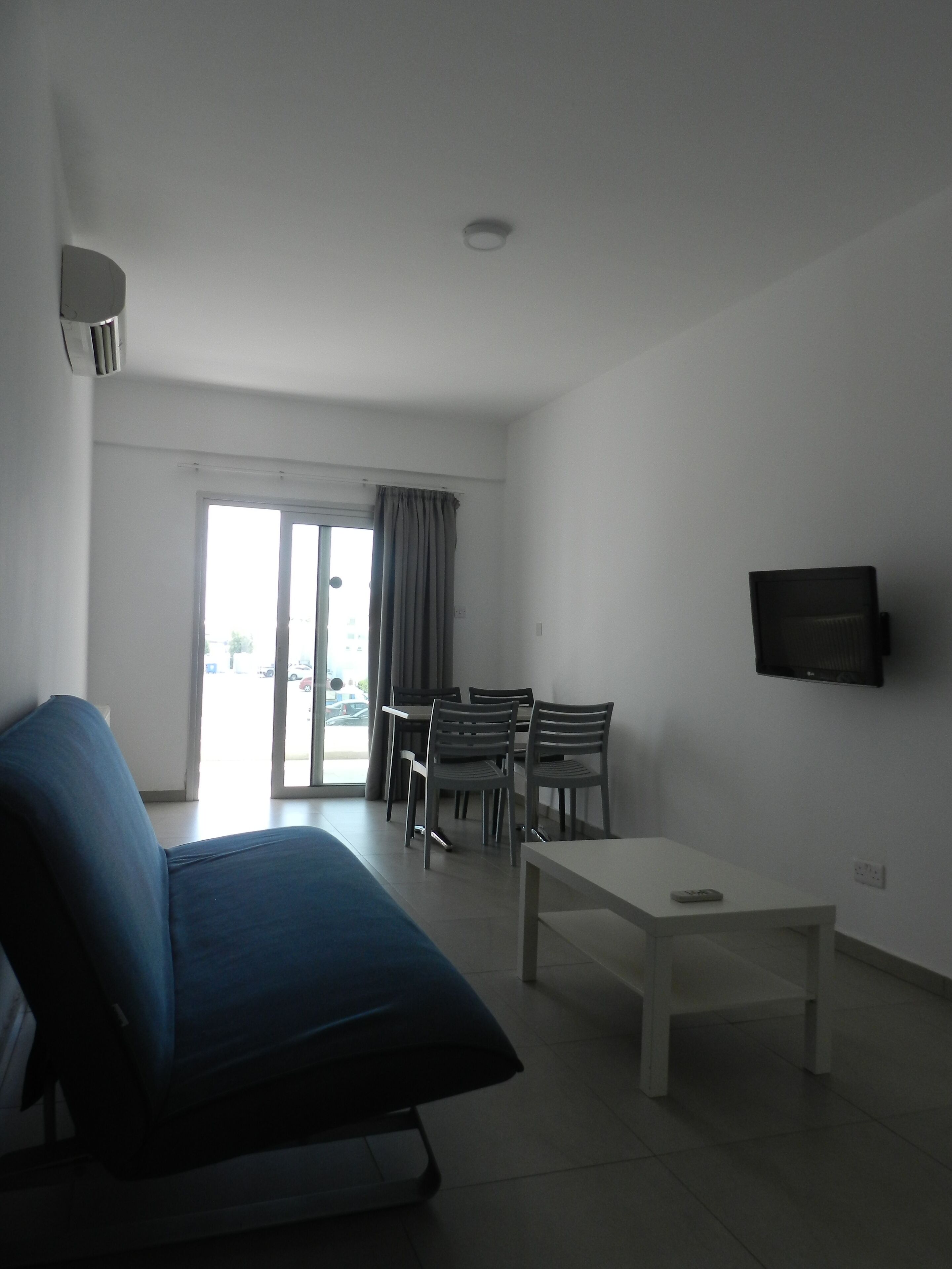 A living room with a sofa, a dining table with chairs, a flat-screen TV mounted on the wall, and a sliding glass door leading to an outdoor area.
