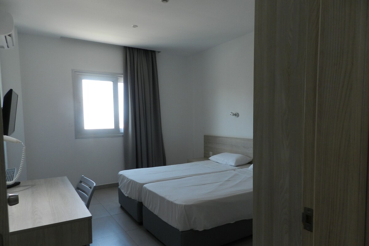 A hotel room with a bed, a desk, a chair, a window with curtains, and a wall-mounted phone.