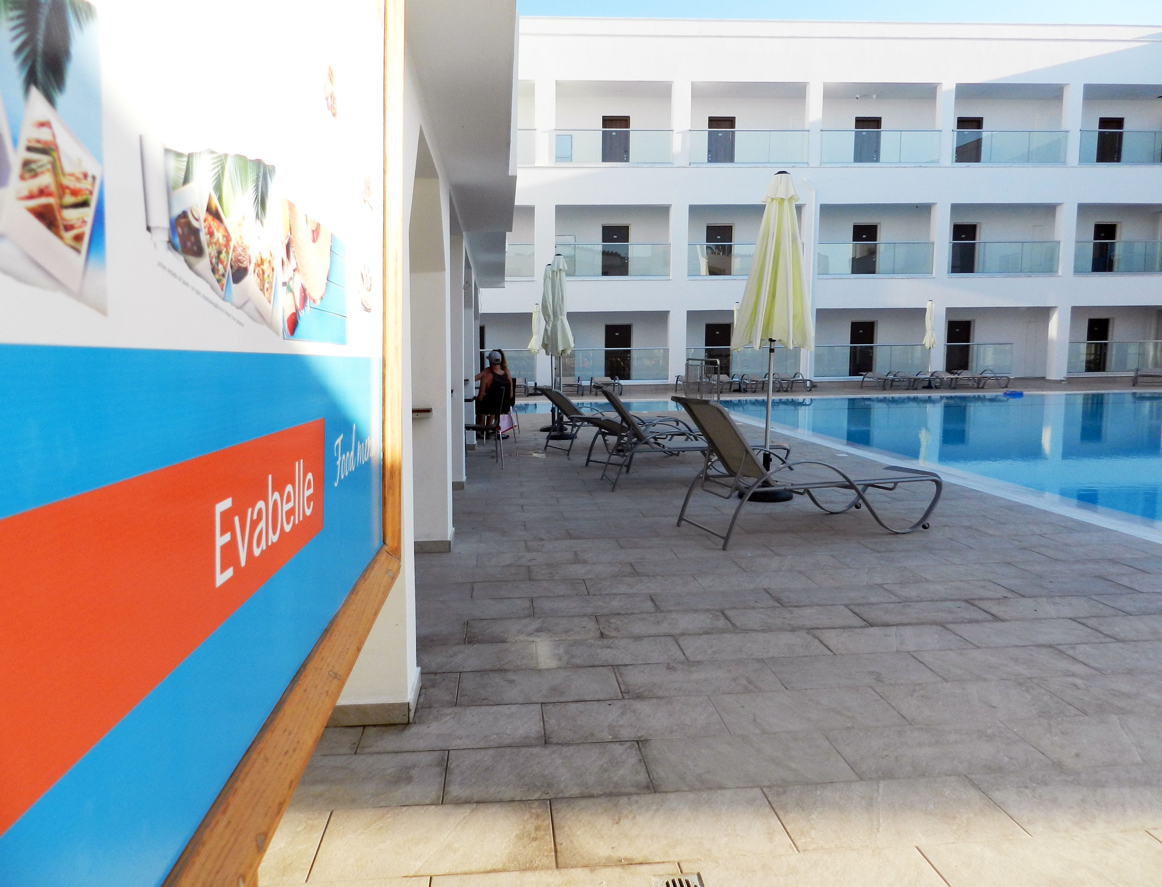 A poolside area with lounge chairs and umbrellas, a pool, and a building with balconies in the background.
