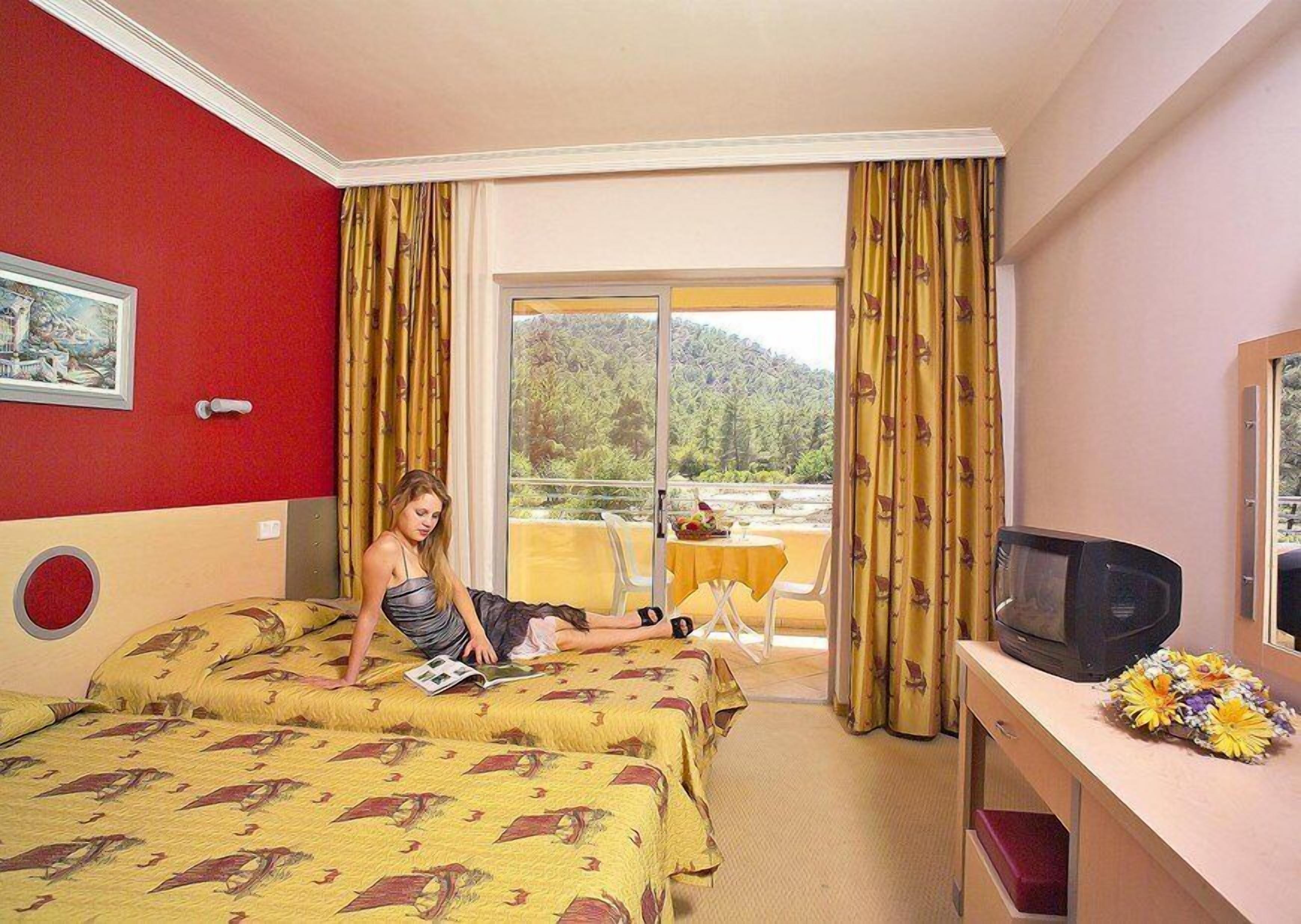 Ein Hotelzimmer mit zwei Betten, einem Fernseher, einem Balkon mit Tisch und einem Gemälde an der Wand.