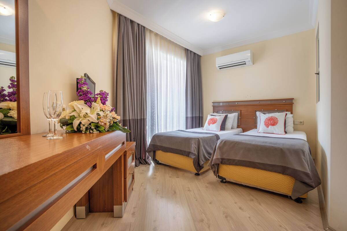 A hotel room with two beds, a wooden desk, a mirror, and a large window with curtains.