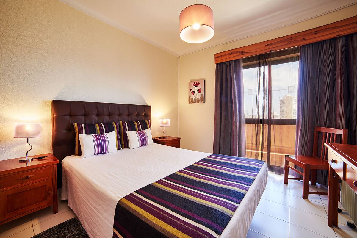 A hotel room with a large bed, a wooden nightstand, a desk, and a window with curtains.