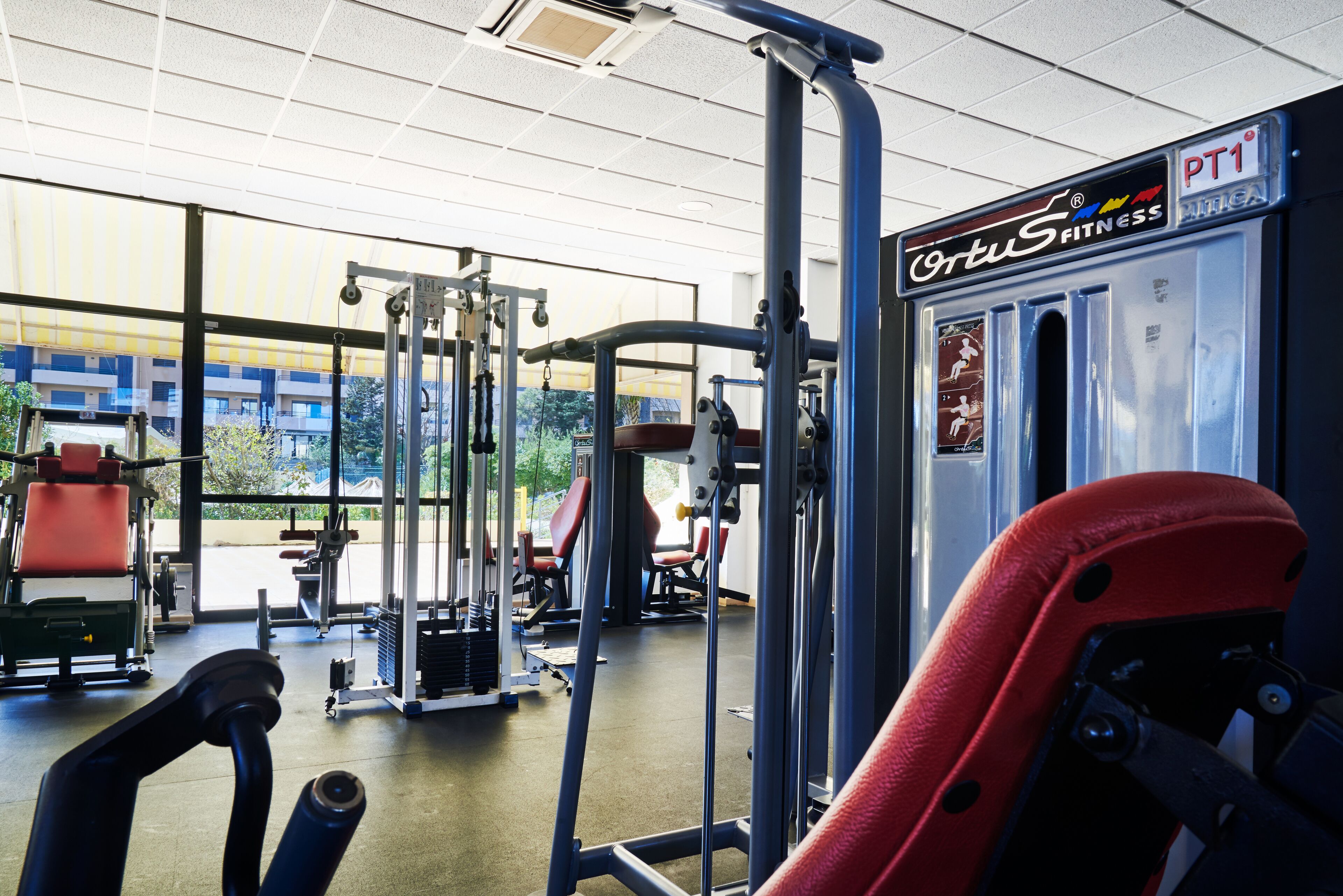 Ett gym med olika träningsmaskiner, en skylt med texten ”Cortus Fitness PT1” och stora fönster som släpper in naturligt ljus.