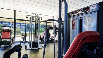 Ett gym med olika träningsmaskiner, en skylt med texten ”Cortus Fitness PT1” och stora fönster som släpper in naturligt ljus.
