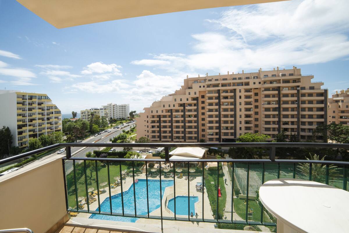 A balcony with a view of a swimming pool, a road, and surrounding buildings.