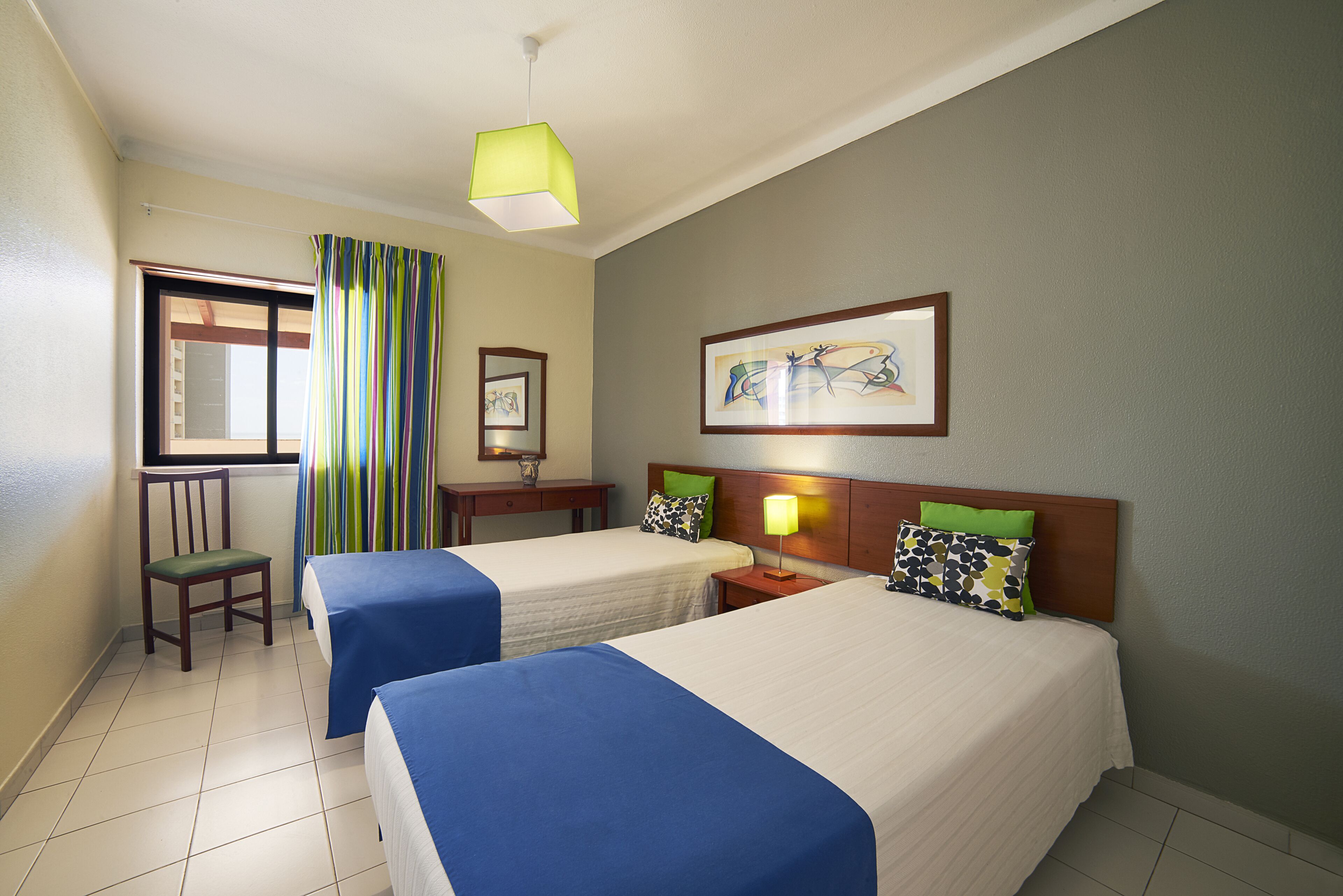 A hotel room with two beds, a chair, a window with curtains, a painting on the wall, and a lamp.