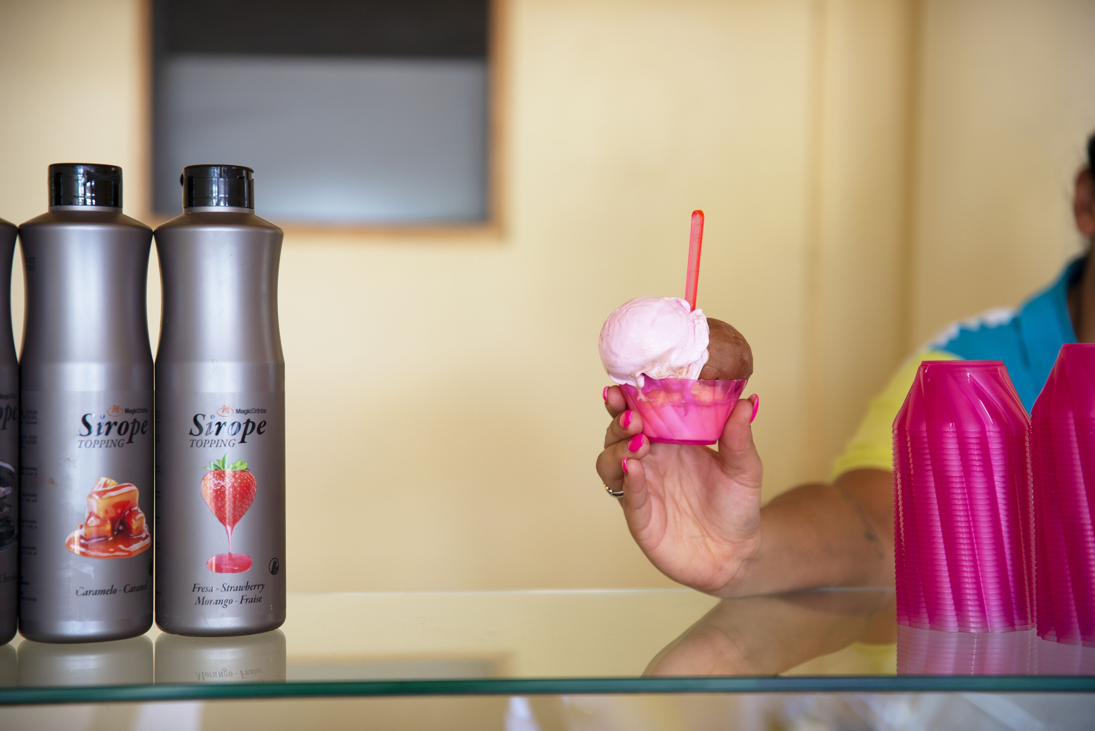 A person holding a pink ice cream cone with a red straw, next to two bottles labeled "Sirope Topping" with images of a strawberry and a caramel.