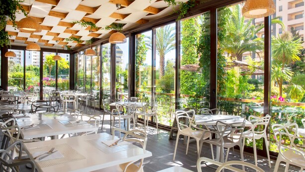 A restaurant with white tables and chairs, large windows with a view of greenery and buildings, and a wooden ceiling with hanging lights. - RR Club  Amarilis (Portimão)