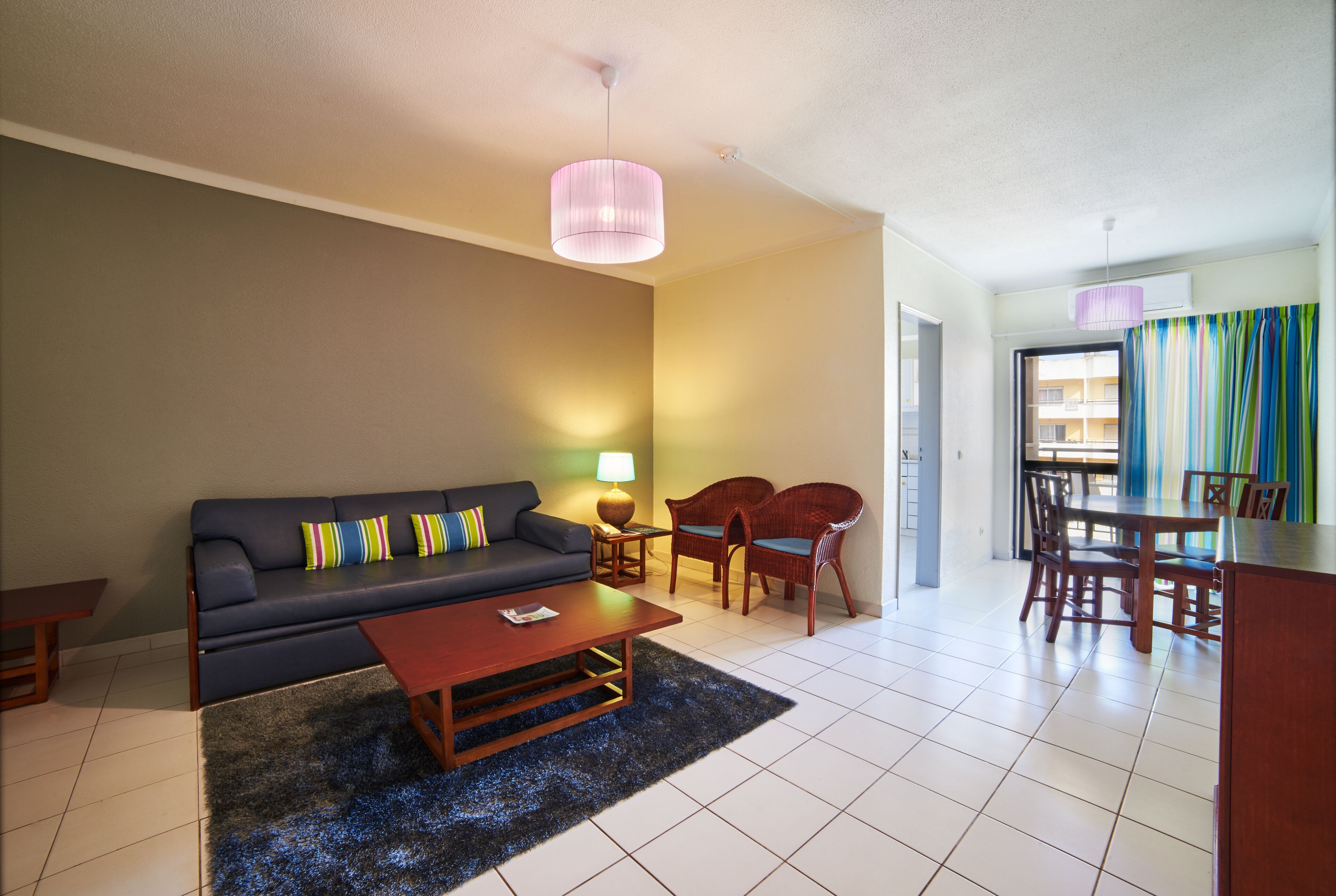 Un salon avec un canapé, une table basse et des fauteuils, un coin repas avec une table et des chaises, et une cuisine visible au fond.
