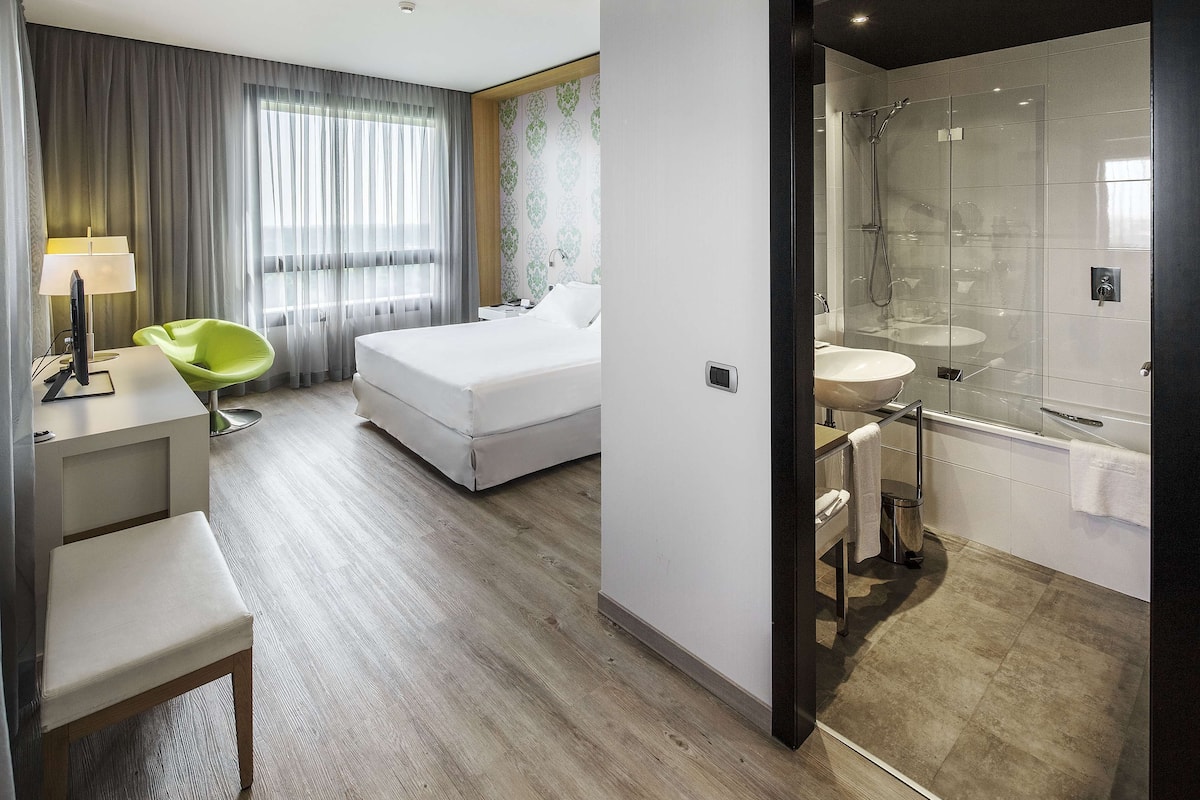 A modern hotel room with a bed, a desk with a lamp, a green chair, and a bathroom with a glass shower enclosure.