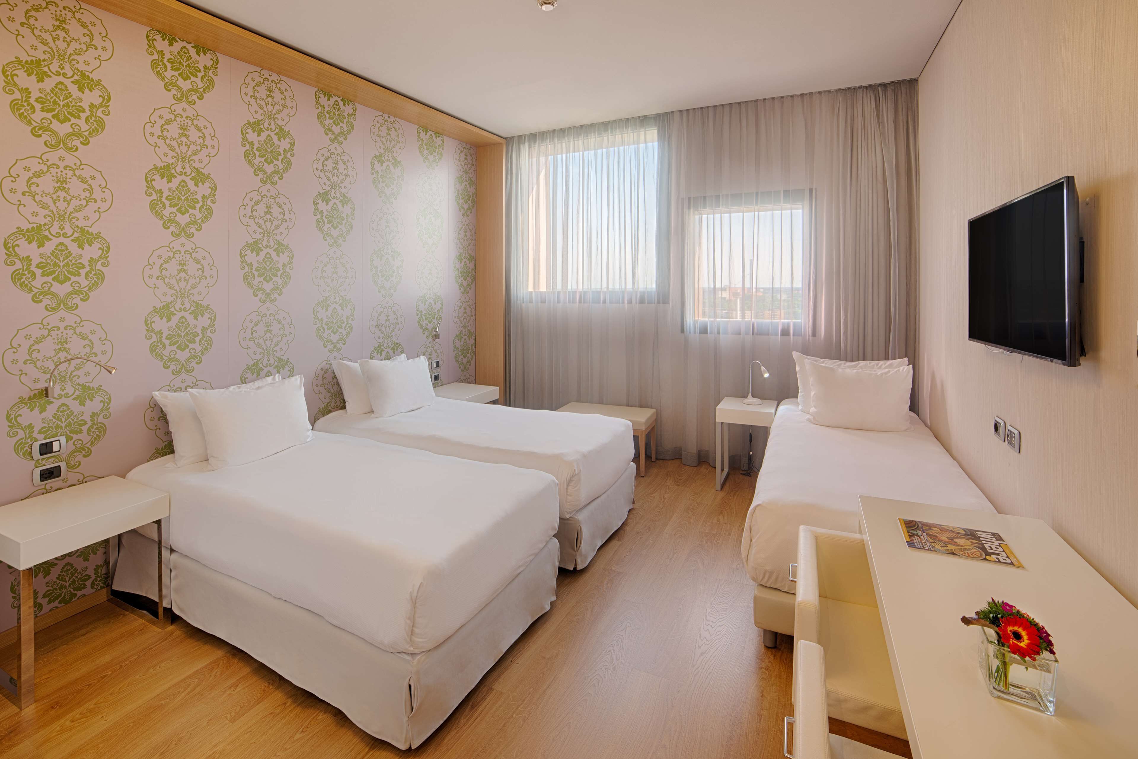 Ein Hotelzimmer mit zwei Betten, einem Fernseher, einem Schreibtisch und einem Fenster mit Vorhängen.