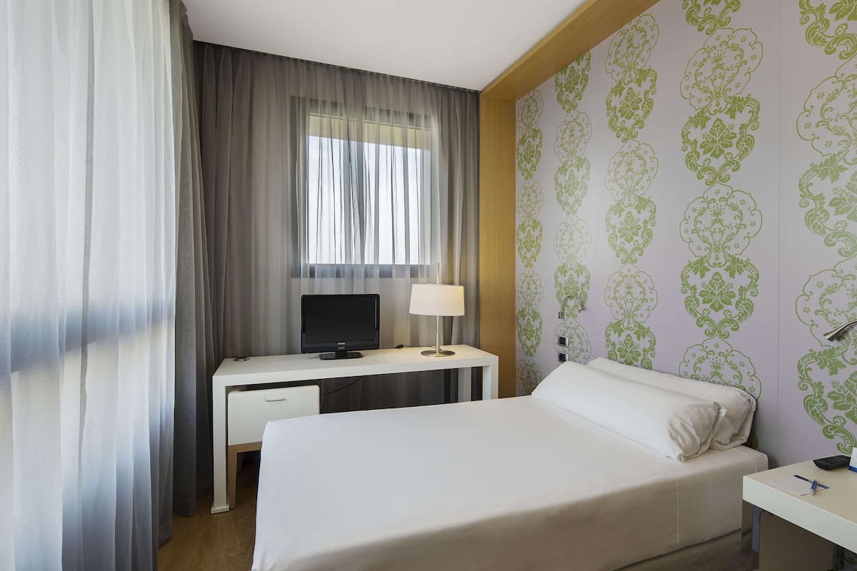 A hotel room with a bed, a desk with a computer, a lamp, and a window with curtains.